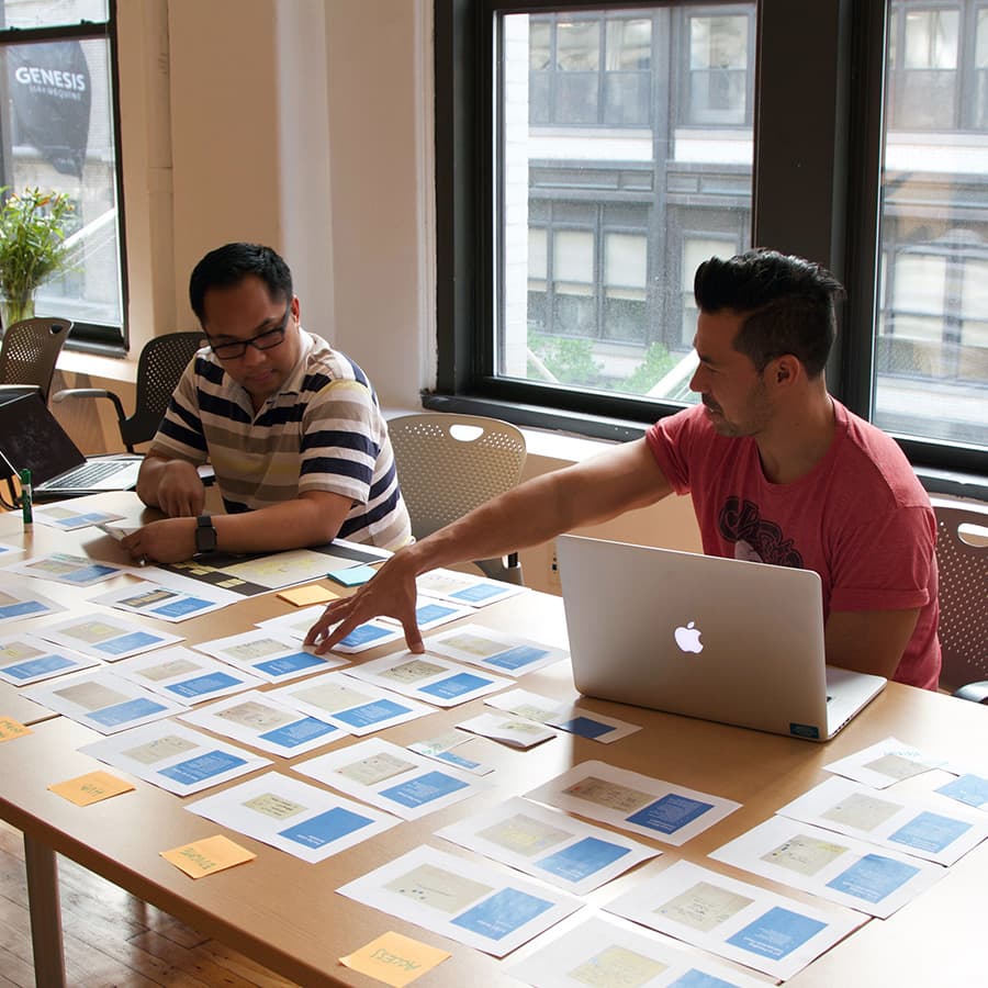 Two men chat at a table with a laptop and sheets of paper lined up with sticky notes on each.